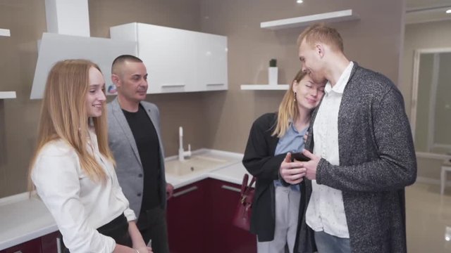 Young Caucasian Couple Standing in Kitchen of New Apartment Property With Real Estate Brokers and Female Recieves Keys While Males Shaking Hands. Light Camera Movement