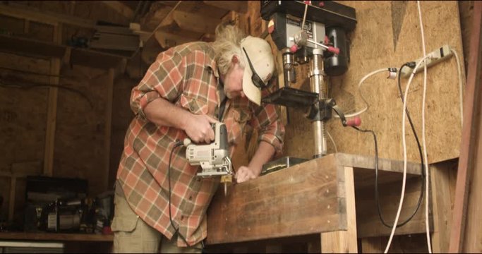 Long-haired carpenter cuts wood with jigsaw in his shop in 4K. Medium wide shot.