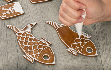 Female hand decorating baked gingerbreads in fish shape. Close-up of painting traditional Christmas sweets by icing bag with white frosting. Cute sweet Xmas cookies with aromatic scent on wood table.