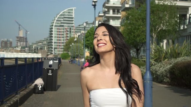 Slow Motion of an attractive and playful latina woman with black wavy hair walking on the banks of the Thames river in London, looking at the camera, spinning, happy with a beautiful smile.