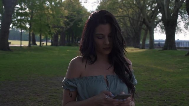 Portrait of a confident and attractive young brunette woman, uses her cellphone, smiles happily to the text messages, looks around. Stylish outfit, modern millennial in slow motion.
