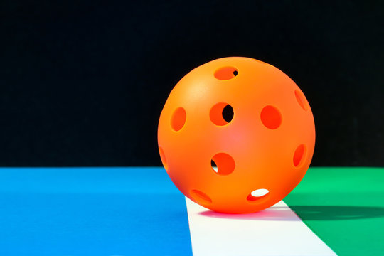 Orange Indoor Pickleball On The Boundary Line Of Pickleball Court With Blue Play Area And Green Out Of Play Area...with A Black Background.