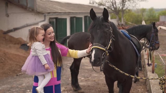 Young Caucasian Mother Holding Her Baby Girl By Black Horses Outside of Stable Barn and They Touching Closer One. Light Camera Movement Blurred Background
