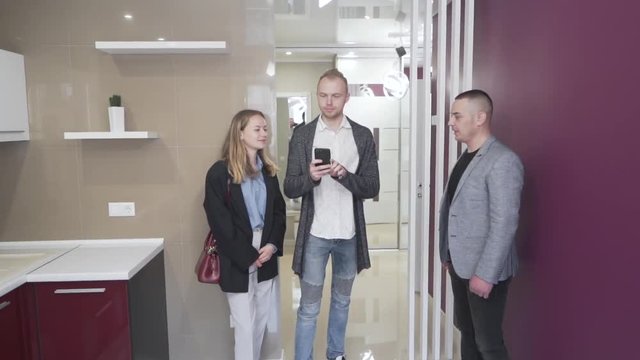 The splendid coule talks with the real estate agent in the modern kitchen. They all observe new apartment that they would like to buy. Tall guy raises his hand with the phone and tries to take photos