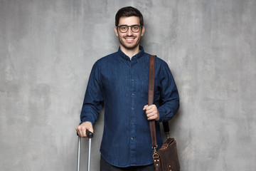 Young smiling modern business man with suitcase and bag ready for coming trip, isolated on textured wall background