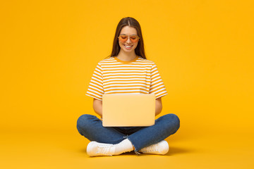 Smiling girl sitting on floor, using laptop, isolated on yellow background