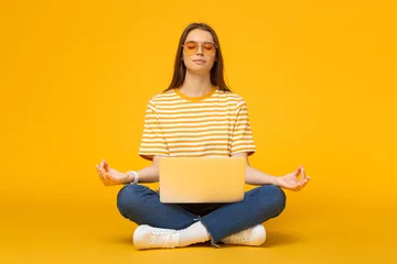 Foto auf Leinwand Junge Frau sitzt auf dem Boden mit Laptop und meditiert in Yoga-Lotus-Pose isoliert auf gelbem Hintergrund © Damir Khabirov