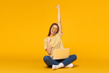 Studio portrait of joyful girl with laptop computer, sitting on a floor and celebrating, isolated...