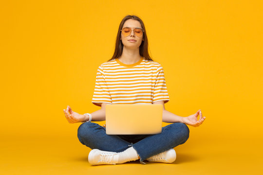 Young woman sitting on floor with laptop meditating in yoga lotus pose isolated on yellow background
