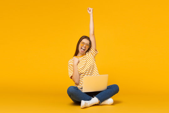 Studio portrait of joyful girl with laptop computer, sitting on a floor and celebrating, isolated on yellow background