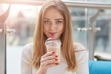 Indoor shot of beautiful young woman drinking milk cocktail, looking directly at camera, hold cup with fresh baverage in her hand, enjoying sweet taste of drinking, wearing white casual shirt.