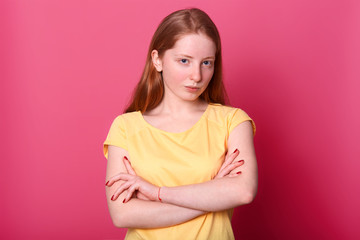 Obraz premium Studio shot of serious harsh girl looking questioningly directly at camera, keeps arms crossed, dressed yellow casual t shirt, isolated over pink background. Copy space for advertisment or promotion.
