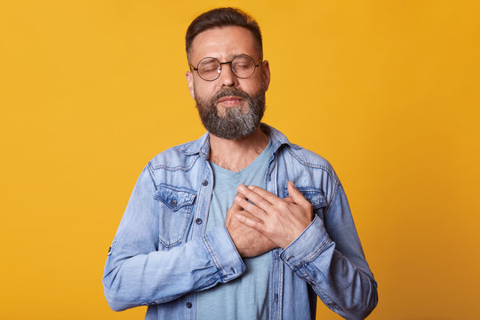 Pleased bearded man with closed eyes, keeps hands on chest, dressed in fashionable denim jacket, isolated over yellow studio background, expresses his love and grateful. People emotions concept.