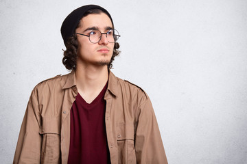 Waist up shot of attractive guy with curly hair, looking aside, wearing hat, fashionable round eyeglasses. red sweatshirt, beige jacket, having pensive facial expression. Copy space for advertisement.