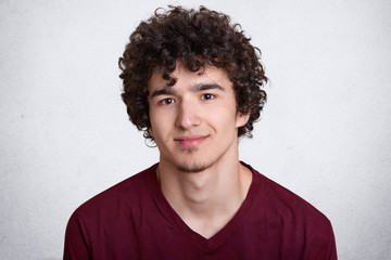 Fototapeta premium Close up portrait of sweet attractive smiling guy with curly hair and beard, having pleasant facial expression, posing isolated over white background in studio, wearing casual red sweatshirt.