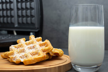 Carrot waffles sprinkled with powdered sugar on a wooden plate with a glass of milk. Waffle iron in the background