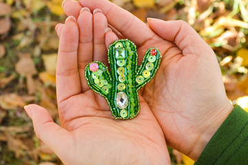 Green cactus toy, handmade bead brooch, lying on women's palms. The background is blurred.