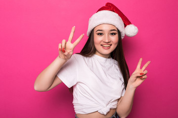 Fototapeta na wymiar Young asian woman wearing christmas hat smiling with happy face winking at the camera doing victory sign isolated over pnk background