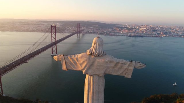 Aeria view monument Sanctuary of Christ the King. Drone flyby past near giant stone sculpture overlooking city of Lisbon Almada and famous 25th april bridge over river Tagus at sunset