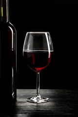 Bottle of red wine and a glass half filled with red wine, on a wooden black table, black background.