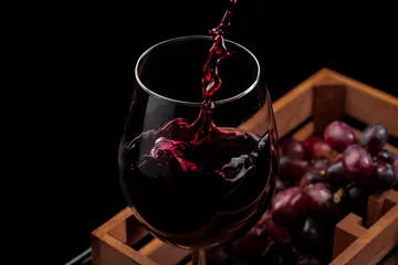 Foto auf Alu-Dibond Beautiful red wine splash in a glass goblet in a wooden box with grapes, black background. © igorgeiger