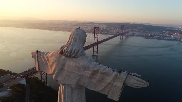 Aeria view monument Sanctuary of Christ the King. Drone flyby past near giant sculpture overlooking city of Lisbon Almada and famous 25th april bridge over river Tagus at sunset