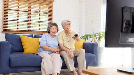 Senior asian couple holding remote control while watching television in home living room with happiness, old people retirement routine lifestyle