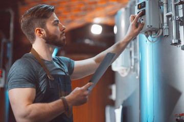 A young handsome brewer in an apron with a tablet in his hands amid the vats of a brewery