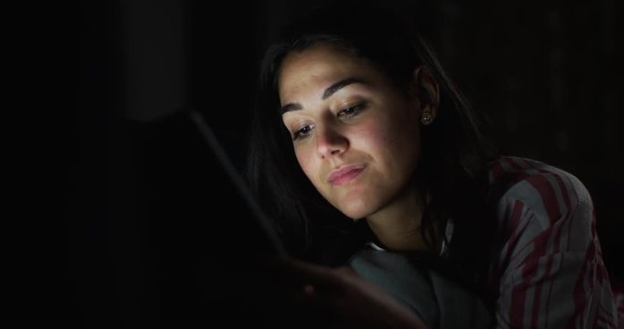 4K Young Hispanic woman lying in the dark & looking at tablet computer. Slow motion.