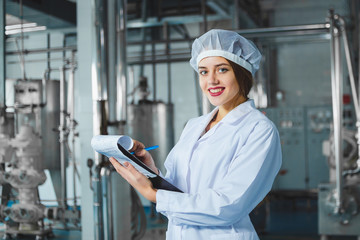 A young beautiful girl in white overalls makes notes in a tablet on the background of equipment of a food processing plant. Quality control in production