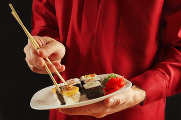 Hands of man in a red shirt are holding a plate with sushi set on a black background