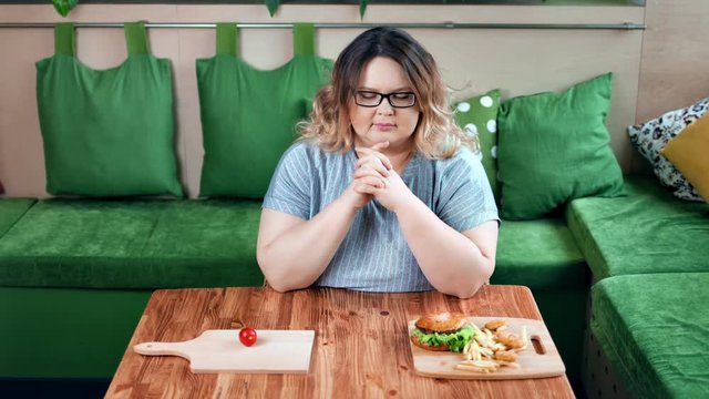 Thoughtful dieting fat woman choosing between American fast food and fresh organic vegetable