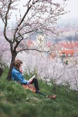 Stylish woman sitting on green grass under blossoming trees and reading a bookon hill in center of Prague, Czech Republic