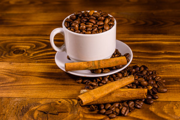White cup filled with coffee beans and cinnamon sticks on wooden table