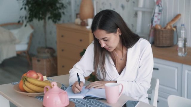 Young woman in bathrobe sits on kitchen and makes notes in notebook, slow motion