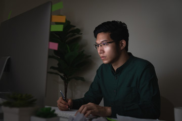 Attractive young asian man sitting on desk table looking at laptop computer in dark late night working feeling serious thinking and determinated at home office in work hard or work load job concept.