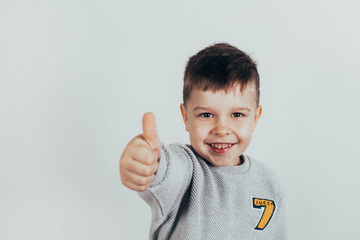Happy child, little boy showing thumbs up gesture in gray sweater against gray background. Space for your text.