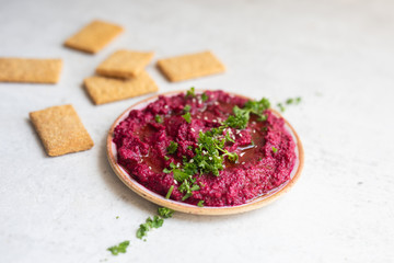 Close up of beet hummus served with hemp seeds, chopped parsley, crackers and a drizzle of olive oil in a pottery bowl on a light natural stone background. Copy space, side view.