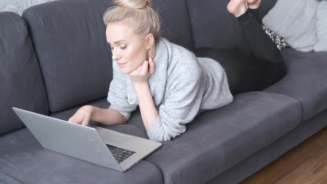 Happy blond woman lying prone on sofa and working on laptop computer