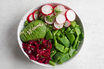 Healthy Buddha Bowl salad with flat green beans, pink radishes, half avocado sliced, shredded cabbage and beets or beetroots, hemp seeds, parsley with vinaigrette in a white pottery bowl. Top view.