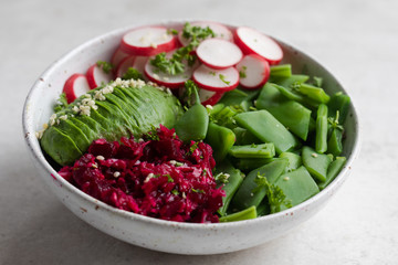 Close up healthy Buddha Bowl salad with flat green beans, pink radishes, half avocado sliced, shredded cabbage and beets or beetroots, hemp seeds, parsley with vinaigrette in a white pottery bowl.