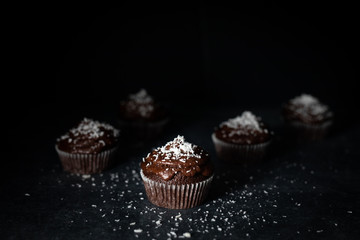 Chocolate cupcakes decorated with a chocolate cream frosting with coconut powder on top and chocolate shavings on a rustic wooden board on a dark background.