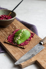 Seed cracker with avocado slices, beet hummus and hemp seeds on a rustic wooden board served with extra beet hummus in a pottery bowl with a rustic knife and a purple napkin. Close up, copy space. 