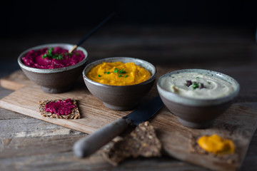 Three hummus dips, beet hummus, sweet potato or carrot or pumpkin hummus and parsnip hummus on a rustic wooden board with a rustic knife on a dark wooden table. Side view