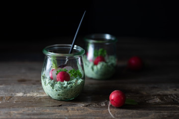 Close up of jars with radish leaves, mint and ricotta dip with fresh radishes, chives and a spoon on a rustic wooden table. Side view, copy space
