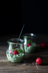 Close up of jars with radish leaves, mint and ricotta dip with fresh radishes, chives and a spoon on a rustic wooden table. Side view, copy space
