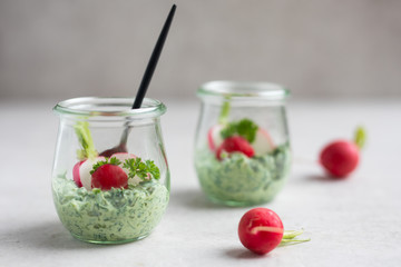 Close up of jar with radish leaves, mint and ricotta dip with fresh radishes, chives and a spoon on a light natural stone table. Side view, copy space