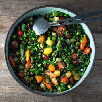 Du Puy lentils, cherry tomatoes, asparagus, sweet peas, scallions, parsley, cilantro, basil salad with vinaigrette in a pottery salad bowl. Top view, rustic wooden background, square. 
