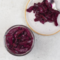 Red pickled/preserved/fermented marinated beets in a Bonne-Maman glass jar with a spoon and a plate on a natural stone light background. Top view and copy space. 