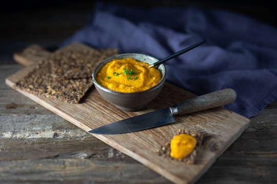 Sweet potato or pumpkin or carrot hummus with parsley and olive oil, served with seed crackers on a rustic wooden board with a spoon. Dark background, side view, copy space.